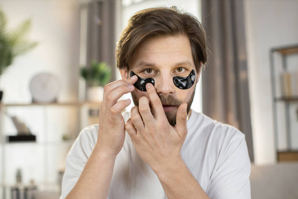 Handsome smiling happy caring young bearded guy poses at camera with anti-aging hydrogel eye-patches - Foto, Bild