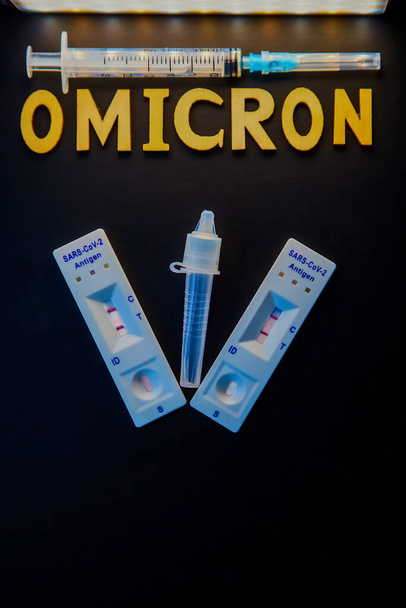 word omicron made from wooden letters,syringe, medical injection bottle and 2 covid-19 antigen self-tests positive on black background.concept-Mutated coronavirus SARS-CoV-2 flu disease pandemic - Photo, Image