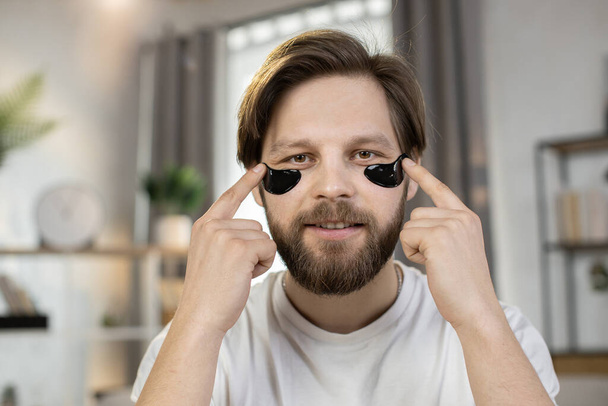 Caucasian man, touching black hydrogel patches under eyes and looking into camera - Zdjęcie, obraz
