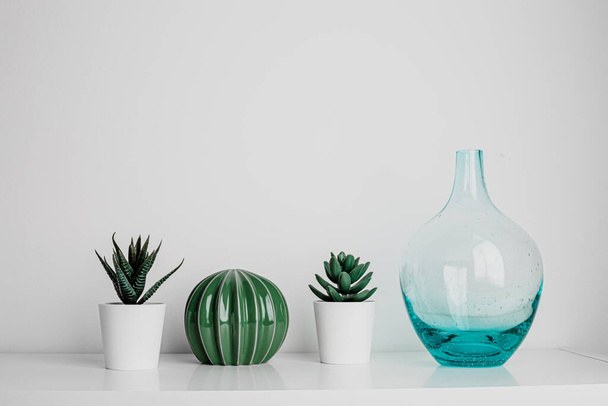 Decorative artificial ceramic and metal cactus, succulent and cactuses on white wall background. Light blue and mint glass vase. Home decoration. Side view. Copy space. - 写真・画像