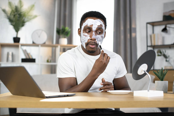 handsome african american applying white clay mask on his face with brush - Fotografie, Obrázek