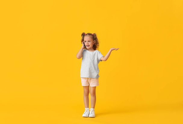 Adorable niña hablando por teléfono móvil sobre fondo amarillo - Foto, Imagen