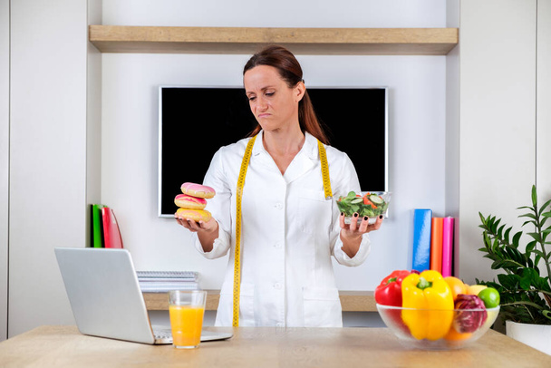Jeune femme portant un uniforme nutritionniste tenant une salade et des beignets avec des émotions tristes dans la salle de bureau moderne lumineux. Choisir entre une alimentation saine et un dessert malsain - Photo, image