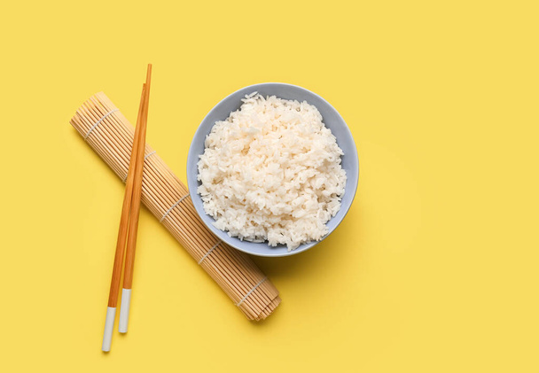 Schüssel mit leckerem gekochtem Reis, Stäbchen und Matte auf gelbem Hintergrund - Foto, Bild