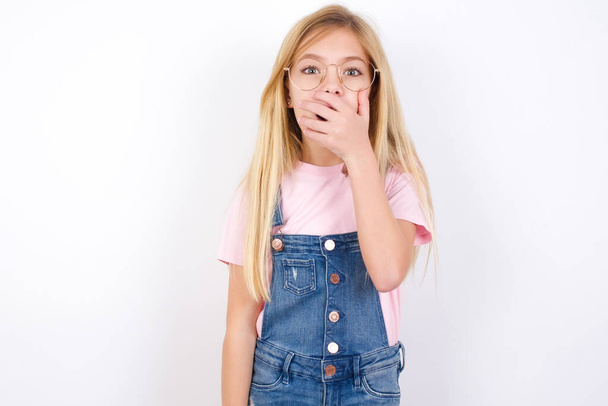 Emotional beautiful caucasian little girl wearing denim jeans overall over white background gasps from astonishment, covers opened mouth with palm, looks shocked at camera. Reaction concept - Fotoğraf, Görsel