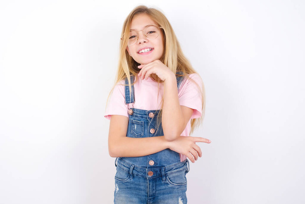 Αισιόδοξη όμορφη καυκάσιος κοριτσάκι φορώντας τζιν συνολικά πάνω από λευκό φόντο κρατά τα χέρια εν μέρει σταυρωμένα και το χέρι κάτω από το πηγούνι, κοιτάζει κάμερα με ευχαρίστηση. Έννοια ευτυχισμένων συναισθημάτων. - Φωτογραφία, εικόνα