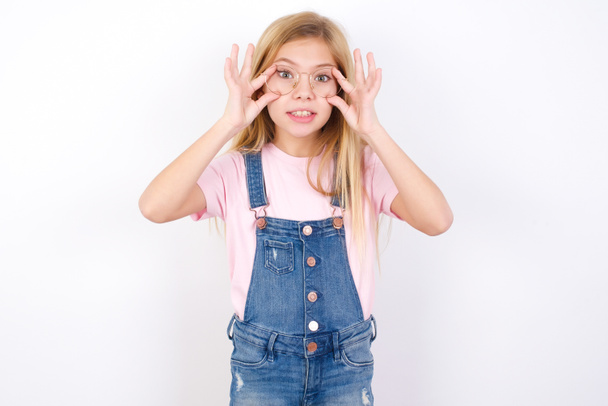 beautiful caucasian little girl wearing denim jeans overall over white background keeping eyes opened to find a success opportunity. - Photo, Image