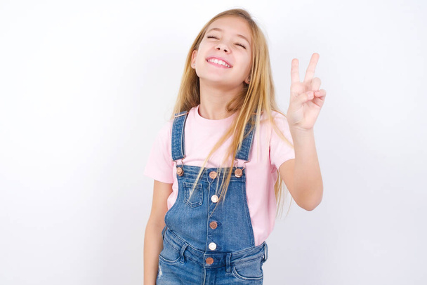 красивая кавказская маленькая девочка в джинсах в целом на белом фоне улыбается со счастливым лицом, подмигивая в камеру, делая знак победы. Номер два:. - Фото, изображение