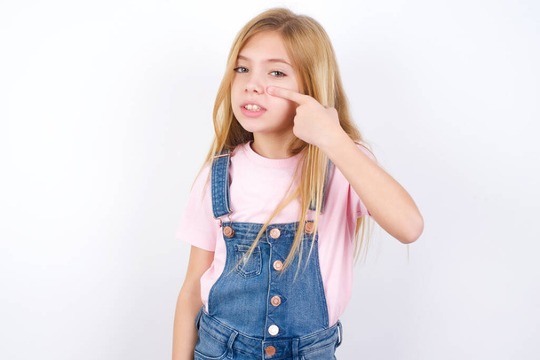 beautiful caucasian little girl wearing denim jeans overall over white background pointing unhappy to pimple on forehead, ugly infection of blackhead. Acne and skin problem - Photo, Image