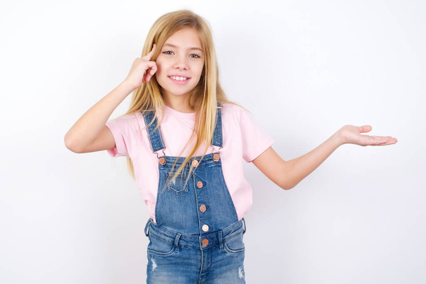 beautiful caucasian little girl wearing denim jeans overall over white background confused and annoyed with open palm showing copy space and pointing finger to forehead. Think about it. - Photo, Image
