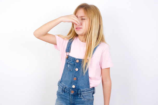 beautiful caucasian little girl wearing denim jeans overall over white background smelling something stinky and disgusting, intolerable smell, holding breath with fingers on nose. Bad smell - Photo, Image