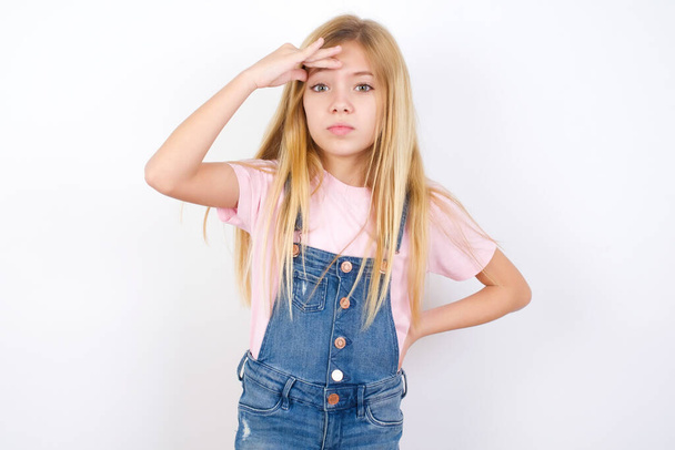 beautiful caucasian little girl wearing denim jeans overall over white background having problems, worried and stressed holds hand on forehead. - Photo, Image