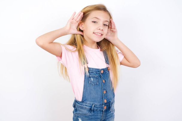 bella bambina caucasica indossa jeans in denim generale su sfondo bianco Cercando di sentire entrambe le mani sul gesto dell'orecchio, curioso di pettegolezzi. Problemi uditivi, sordi - Foto, immagini