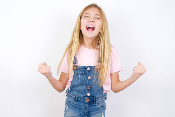 beautiful caucasian little girl wearing denim jeans overall over white background celebrating surprised and amazed for success with arms raised and eyes closed. Winner concept. - Foto, afbeelding