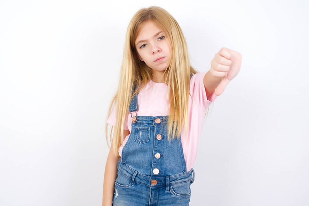 beautiful caucasian little girl wearing denim jeans overall over white background feeling angry, annoyed, disappointed or displeased, showing thumbs down with a serious look - Photo, Image