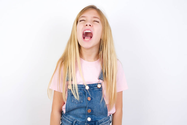 beautiful caucasian little girl wearing denim jeans overall over white background angry and mad screaming frustrated and furious, shouting with anger. Rage and aggressive concept. - Photo, Image