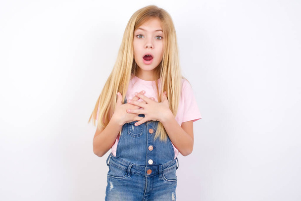 Scared beautiful caucasian little girl wearing denim jeans overall over white background looks with frightened expression, keeps hands on chest, being puzzled to notice something strange, People, hush reaction and emotions. - Фото, изображение
