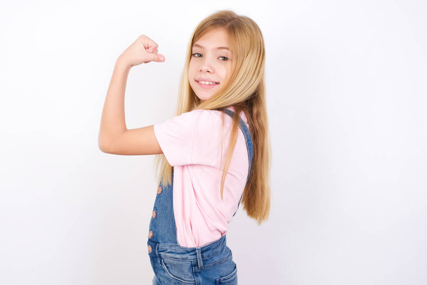 Portrait of powerful cheerful beautiful caucasian little girl wearing denim jeans overall over white background showing muscles. - Photo, Image