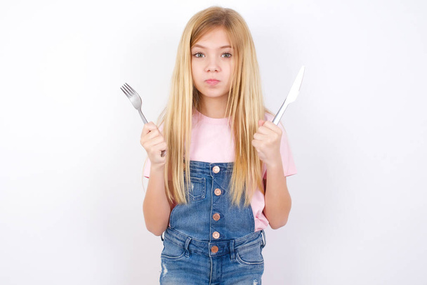Model hongerig mooi Kaukasisch meisje dragen spijkerbroek overall over witte achtergrond houden in de hand vork mes wil lekker lekker pizza taart - Foto, afbeelding
