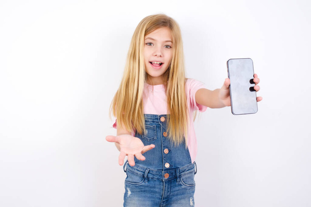 beautiful caucasian little girl wearing denim jeans overall over white background with a mobile. presenting smartphone. Advertisement concept. - Fotografie, Obrázek