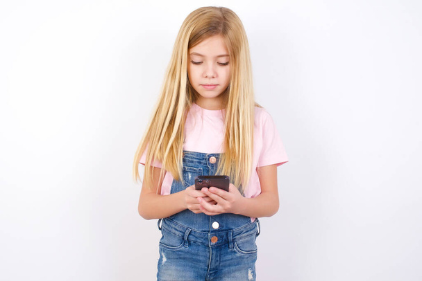 Excited beautiful caucasian little girl wearing denim jeans overall over white background winking and eye hold smart phone use read social network news - Fotó, kép