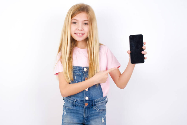 Smiling beautiful caucasian little girl wearing denim jeans overall over white background Mock up copy space. Pointing index finger on mobile phone with blank empty screen - Fotó, kép