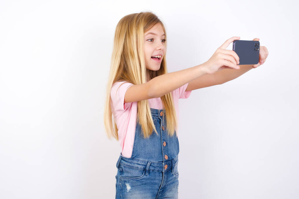 красивая кавказская маленькая девочка в джинсах в целом на белом фоне делает селфи, чтобы разместить его в социальных сетях или иметь видео-звонок с друзьями. - Фото, изображение
