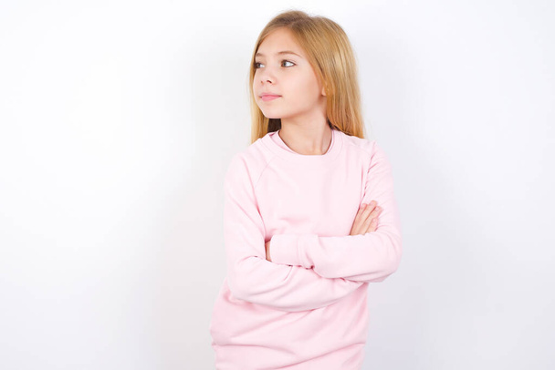 Чарівна красива кавказька дівчинка в рожевому светрі на білому тлі стоїть зі складеними руками, зосередженими десь з пустотливим виразом думає, що робити
 - Фото, зображення