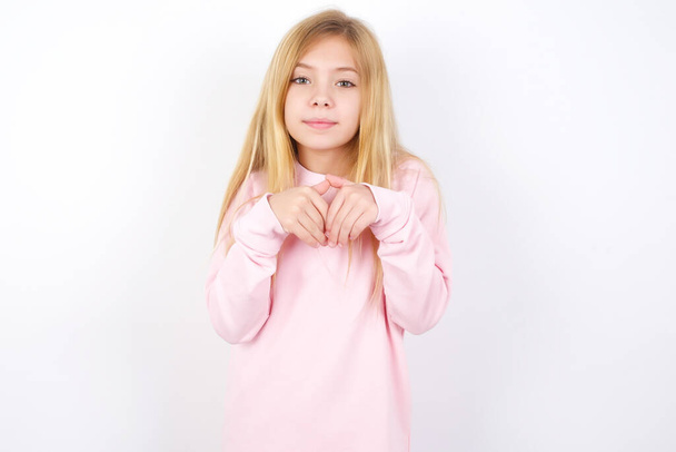 bella caucasica bambina indossa maglione rosa contro sfondo bianco fa zampe di coniglio e guarda con espressione innocente gioca con il suo bambino - Foto, immagini