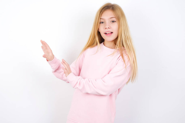 Dispiaciuto bella caucasica bambina indossa maglione rosa contro sfondo bianco tiene le mani verso lo spazio vuoto e chiede di non avvicinarsi vede qualcosa di spiacevole - Foto, immagini