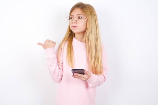 όμορφο καυκάσιο κοριτσάκι φορώντας ροζ πουλόβερ έναντι λευκό φόντο δείχνει τον αντίχειρα μακριά και δείχνει κενό χώρο στην άκρη, κρατά το κινητό τηλέφωνο για την αποστολή μηνυμάτων κειμένου. - Φωτογραφία, εικόνα