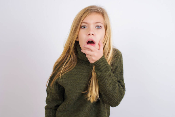 神経質な困惑白人の子供の女の子は白い背景に緑色のニットセーターを着て驚きから口を開き、突然のニュースに反応. - 写真・画像