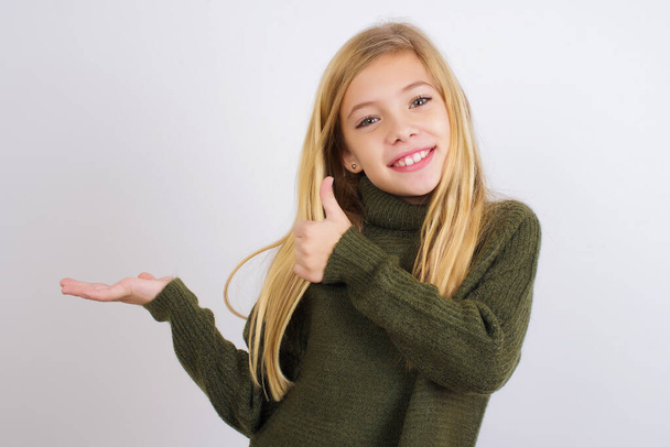 Niña caucásica vistiendo suéter de punto verde sobre fondo blanco mostrando la palma de la mano y haciendo un gesto bien con los pulgares hacia arriba, sonriendo feliz y alegre. - Foto, Imagen