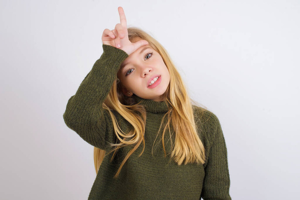 Caucasian kid girl wearing green knitted sweater against white background making fun of people with fingers on forehead doing loser gesture mocking and insulting. - Foto, Imagen