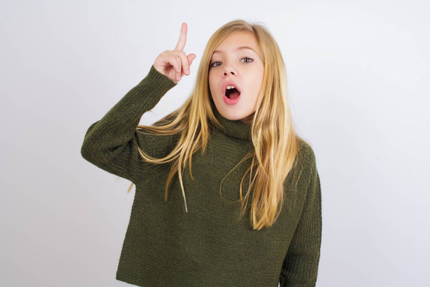 Caucasian kid girl wearing green knitted sweater against white background pointing finger up and looking inspired by genius thought, showing good idea sign, having clever solution in mind - Foto, Bild