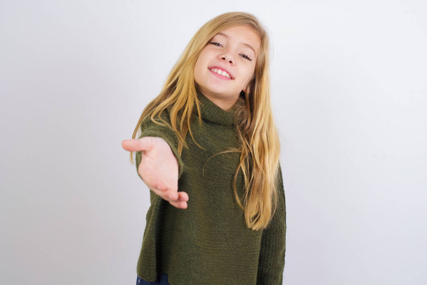 Кавказская девочка в зеленом трикотажном свитере на белом фоне улыбается дружелюбно предлагая рукопожатие в качестве приветствия и приветствия. Успешный бизнес. - Фото, изображение