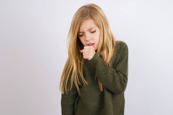 Blank meisje met een groene gebreide trui tegen een witte achtergrond die zich onwel voelt en hoestend als symptoom voor verkoudheid of bronchitis. Gezondheidszorgconcept. - Foto, afbeelding
