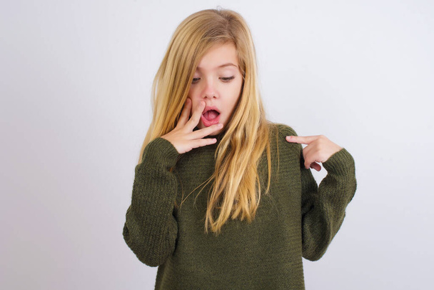 Šokovaný kavkazský kluk dívka na sobě zelené pletený svetr proti bílému pozadí pohled překvapivě dolů, ukazuje na prázdné s předním prstem, Vyděšený model - Fotografie, Obrázek