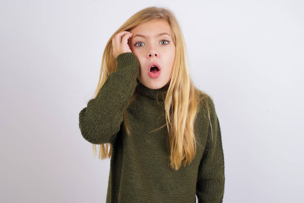 Menina criança caucasiana envergonhada vestindo camisola de malha verde contra fundo branco com expressão chocada, expressa grande espanto, modelo intrigado poses indoor - Foto, Imagem