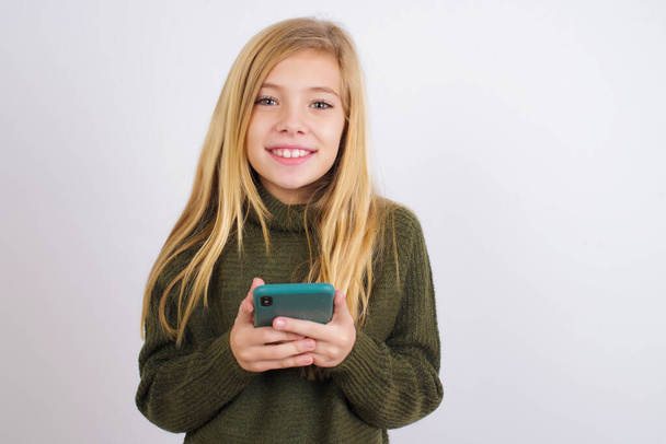  Caucasian kid girl wearing green knitted sweater against white background enjoys distant communication, uses mobile phone, surfs fast unlimited internet, has pleasant smile, makes shopping online, - Foto, Bild