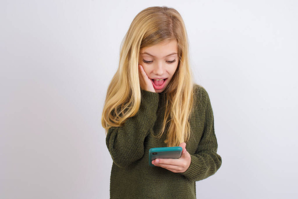 Caucasian kid girl wearing green knitted sweater against white background holding in hands cell reading sms using new app 5g - Φωτογραφία, εικόνα