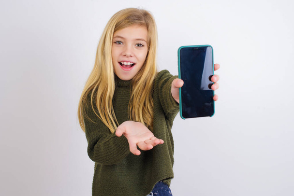 Caucasian kid girl wearing green knitted sweater against white background with a mobile. presenting smartphone. Advertisement concept. - Fotó, kép