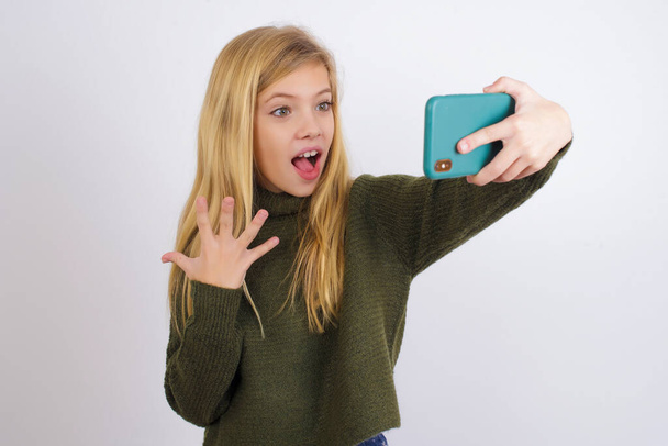 Ouah ! excité enfant caucasien fille portant vert tricoté pull sur fond blanc montrant téléphone mobile avec geste de la main ouverte - Photo, image
