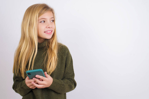 Кавказька дівчинка у зеленому в'язаному светрі на білому фоні утримує телефонні руки і читає хороші новини для молоді виглядають порожніми рекламою простору - Фото, зображення