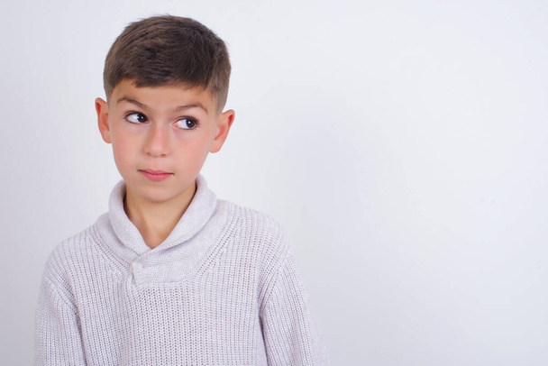 Amazed Caucasian kid boy wearing knitted sweater standing over white background bitting lip and looking tricky to empty space. - Photo, Image