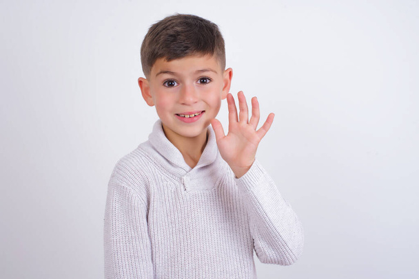 Хлопчик з Кавказу у в'язаному светрі стоїть на білому фоні і вітає з радістю і усмішкою, дружнім жестом.. - Фото, зображення