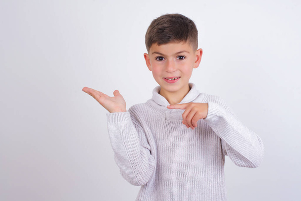 Кавказький хлопчик у в'язаному светрі стоїть над білим фоном і тримає за руку рекламу - Фото, зображення