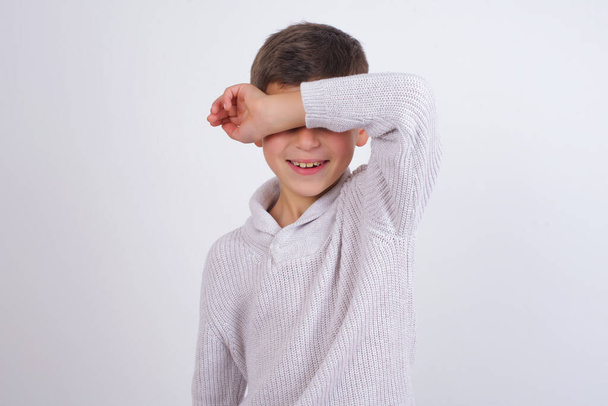 Biały chłopiec noszący sweter na drutach, stojący nad białym tłem, pokrywający oczy uśmiechem na ramieniu i zabawą. Koncepcja niewidomych. - Zdjęcie, obraz