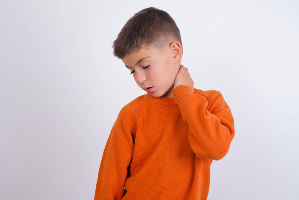 Белый мальчик в вязаном оранжевом свитере, стоящем на белом фоне, страдающий от травмы шеи, трогательной шеи рукой, мышечной боли - Фото, изображение