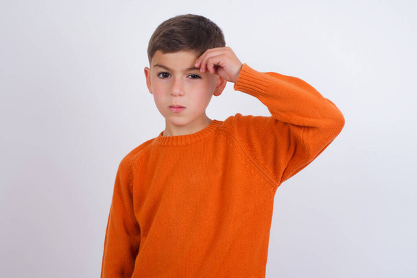 Белый мальчик в вязаном оранжевом свитере стоит на белом фоне, указывая несчастным на прыщ на лбу, заражение угрей головы. Концепция Skincare. - Фото, изображение